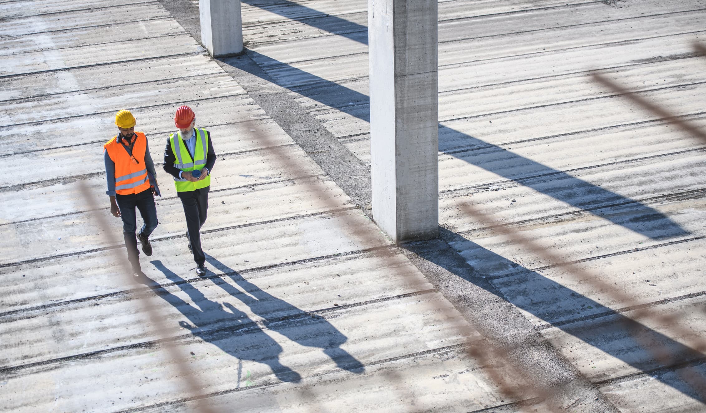 Two people in hardhats and safety vests walk along a wooden surface